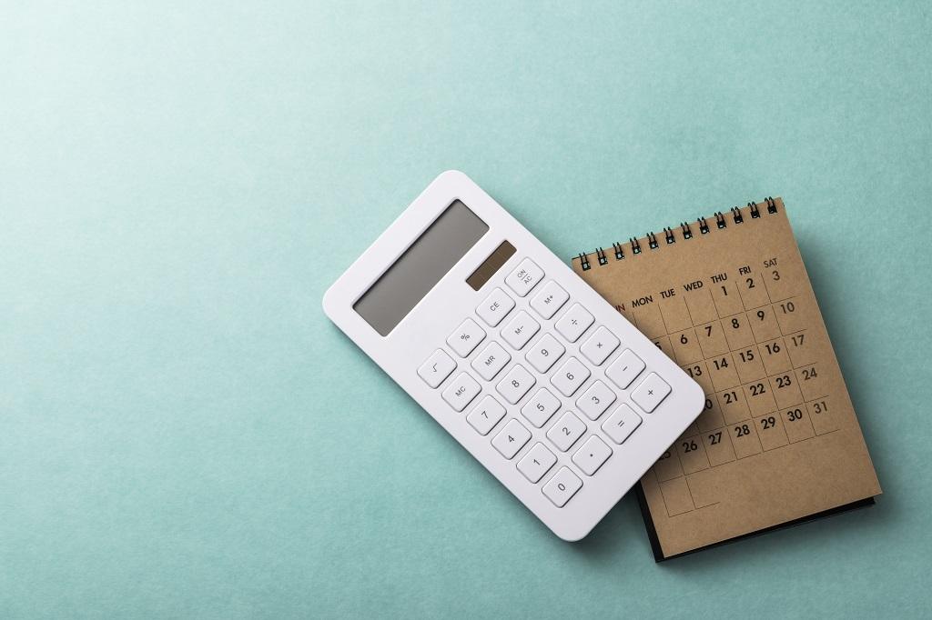 Une calculatrice blanche se trouve à côté d’un petit calendrier marron à reliure spirale sur une surface bleu sarcelle. Le calendrier est ouvert sur une vue mensuelle, affichant les dates et les jours de la semaine. Les éléments suggèrent des thèmes de planification, de budgétisation ou d’organisation.