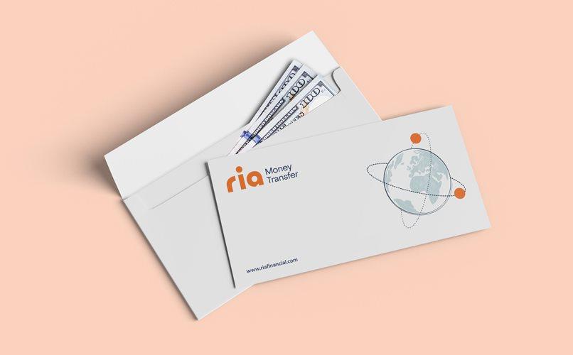 Un sobre con billetes de cien dólares parcialmente visibles se muestra junto a una tarjeta con el logotipo de Ria Money Transfer, que presenta un globo terráqueo abstracto y puntos naranjas. La tarjeta incluye la URL www.riamoneytransfer.com en la parte inferior. El fondo es melocotón suave.