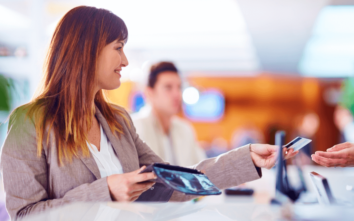 A woman with shoulder-length brown hair and a beige blazer is handing a credit card to an unseen person at a counter. She holds a wallet in her other hand. The background depicts a softly focused setting with posters of Best European Banks, another person, and a bright, modern interior.