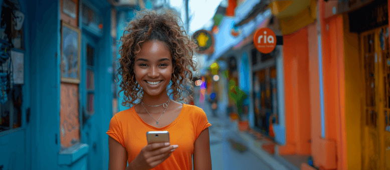 A young woman with curly hair and an orange top smiles while holding a smartphone, standing on a colorful street with blue and orange buildings. The street appears vibrant with signs and decorations in the background.