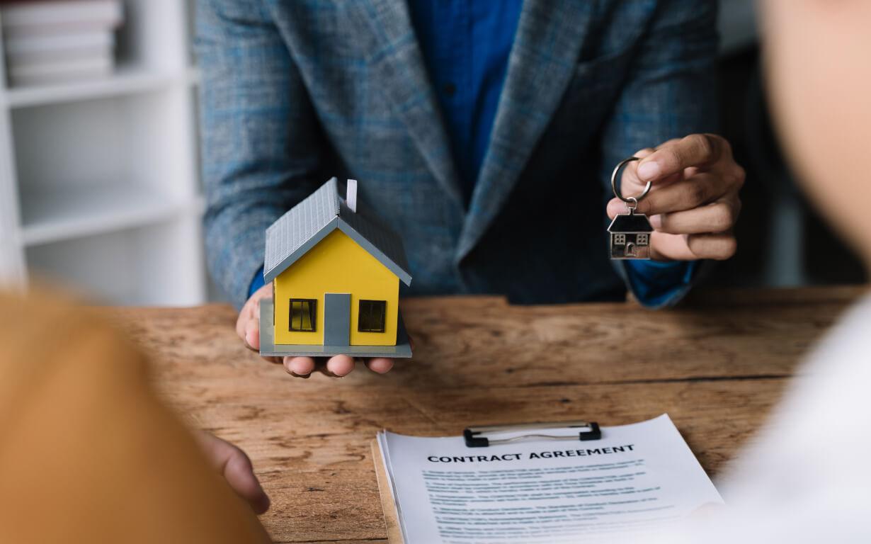 A person in a blue suit holds a small yellow house model and silver keys, facing two other people, indicating they're in the process of buying property. A contract agreement rests on the wooden table before them, suggesting a real estate transaction is underway.