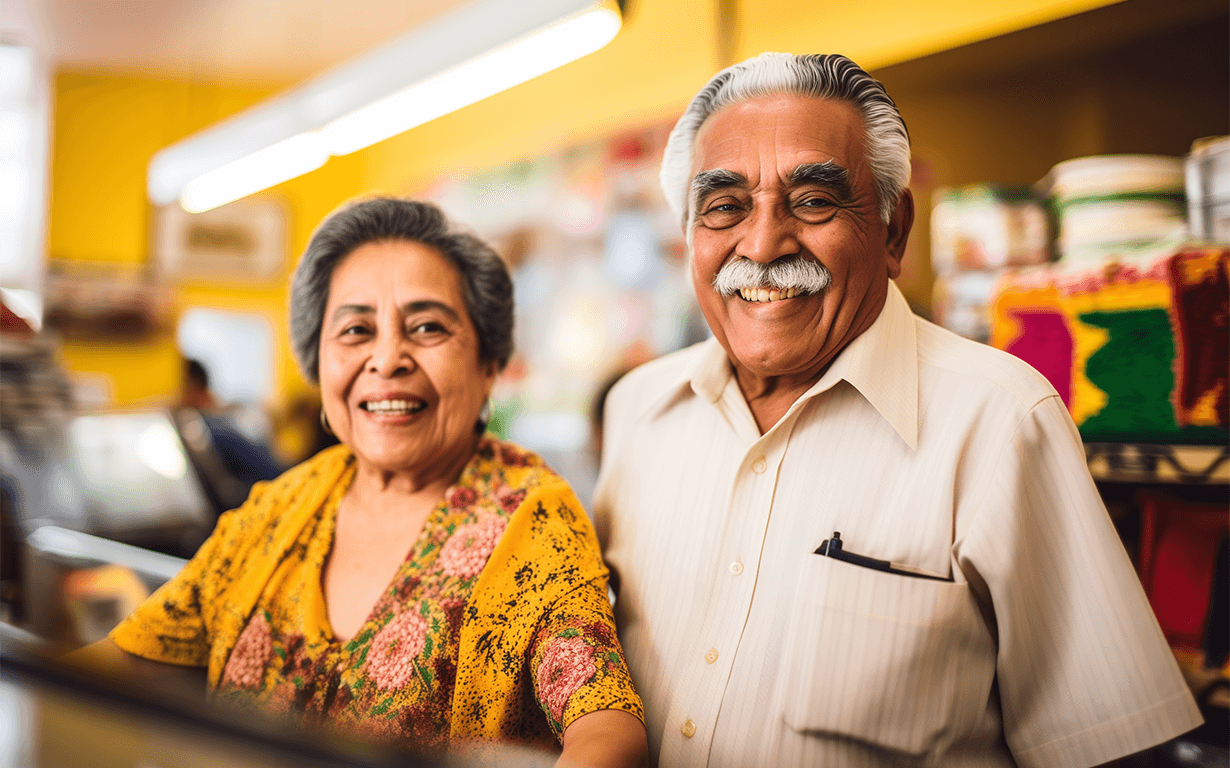 An older couple smiles warmly behind a counter in a brightly lit store with yellow walls and colorful products—offering friendly service and a welcoming spot to send money to Mexico with ease.