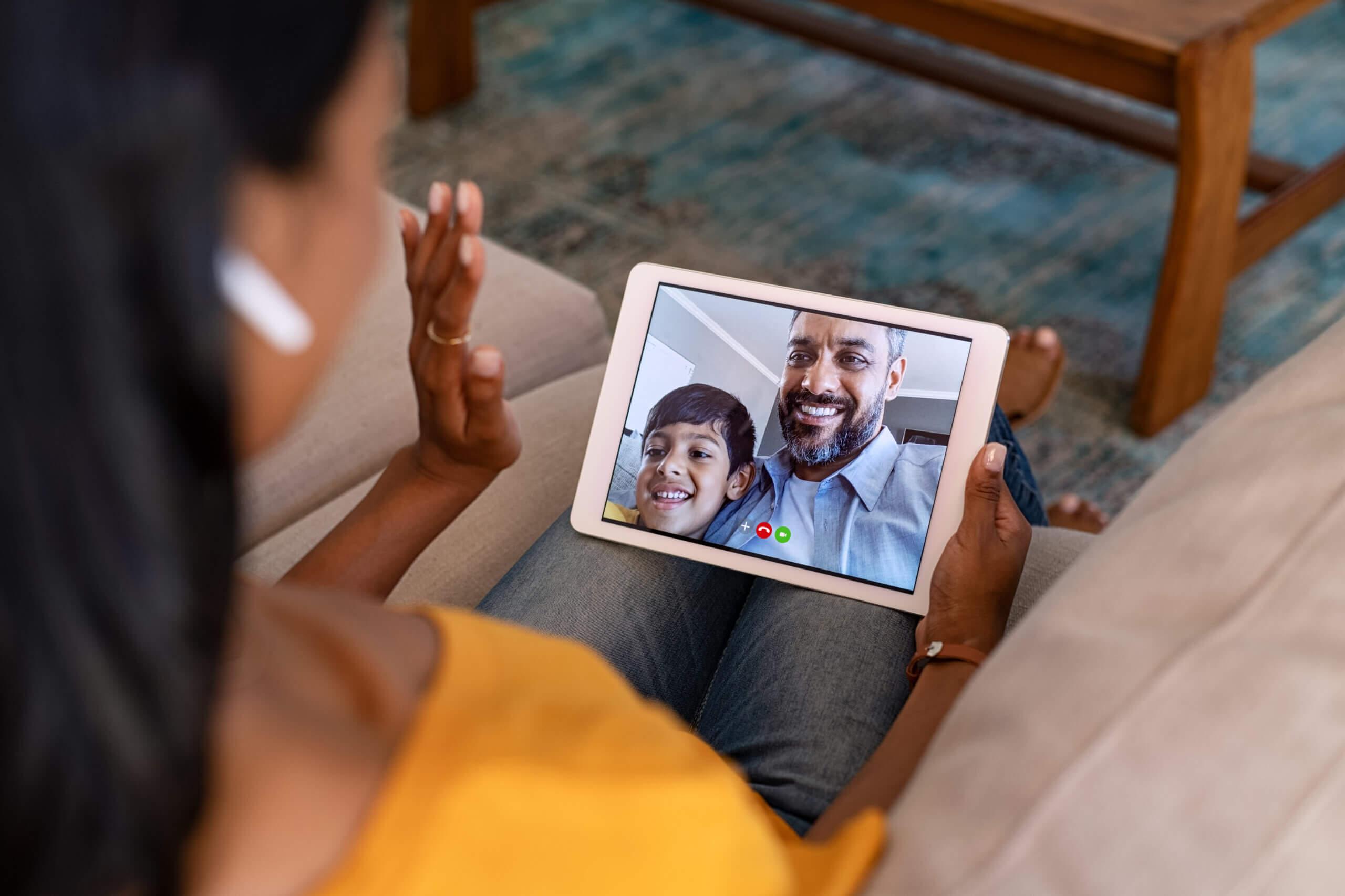 A woman sitting on a couch holds a tablet and video chats with a smiling man and child. She waves at the screen, wearing wireless earbuds. The background shows a rug and a wooden table.